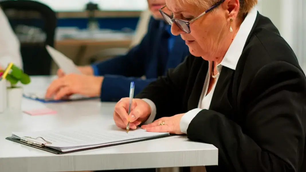 Une femme âgée, vêtue d’une tenue professionnelle, signe un document sur un bureau dans un environnement de travail, tandis qu’une autre personne consulte des papiers en arrière-plan. Représentation d'une mandataire judiciaire.