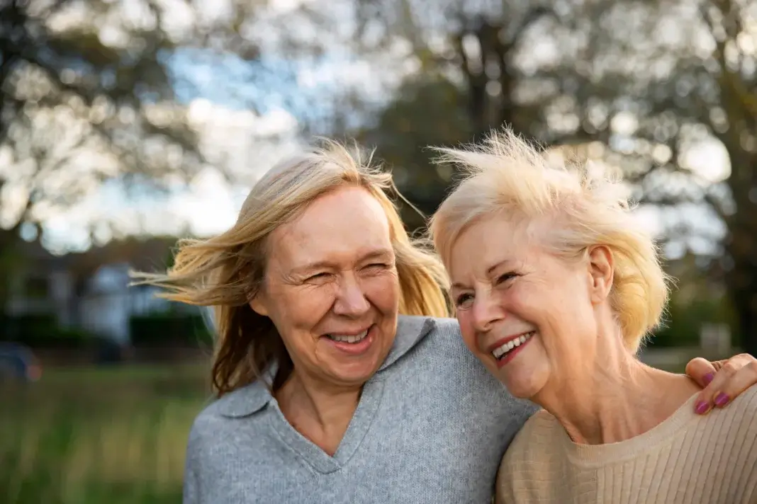 Deux femmes âgées souriantes, proches l’une de l’autre, se tiennent par l’épaule en extérieur, dans un cadre naturel avec des arbres en arrière-plan.