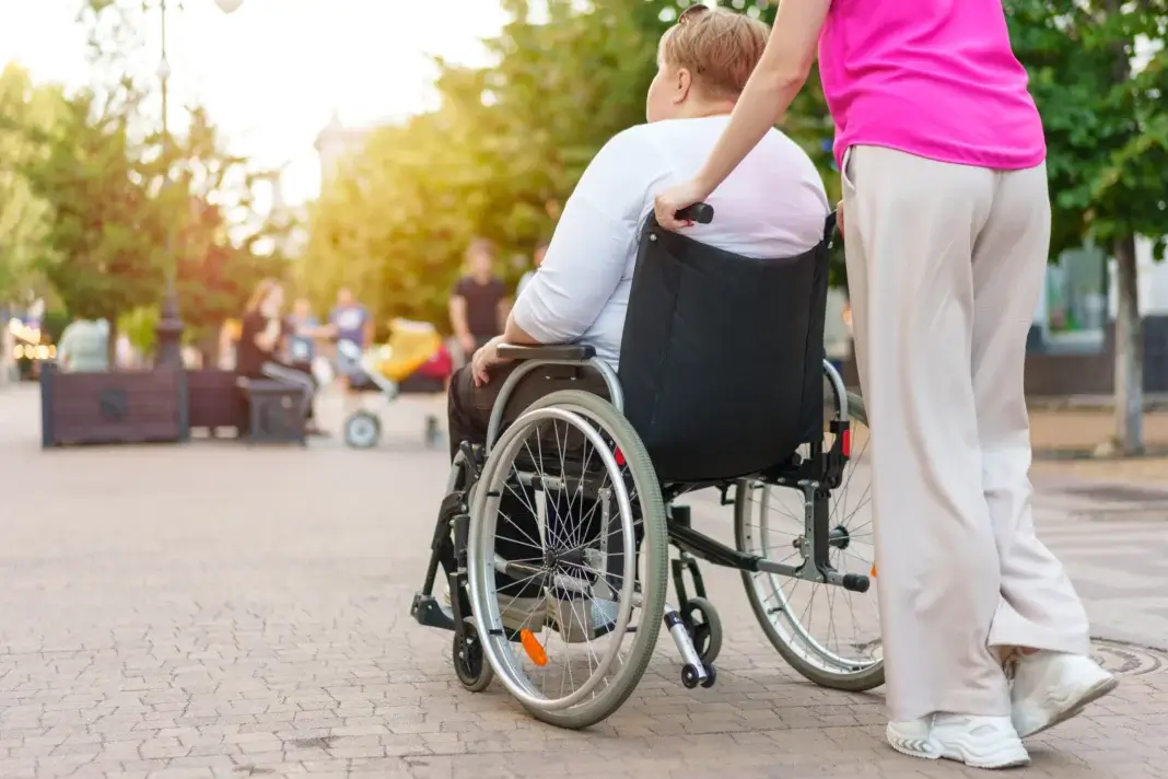 Personne aidante poussant une femme en fauteuil roulant sur un chemin en extérieur, dans un espace public arboré, illustrant le soutien quotidien et la présence des proches aidants auprès des personnes en perte d’autonomie.