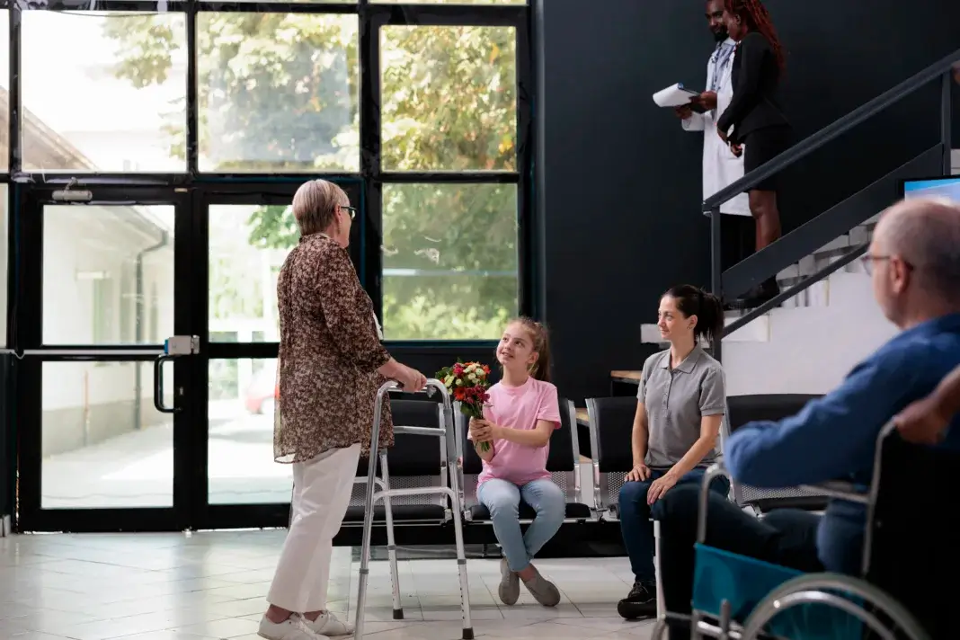 Femme âgée utilisant un déambulateur recevant un bouquet de fleurs d’une jeune fille assise à côté d’une adulte, dans le hall lumineux d’un établissement médico-social, avec d’autres résidents et deux professionnels en arrière-plan près d’un escalier. Image qui illustre une visite par des proches d'une femme âgée.
