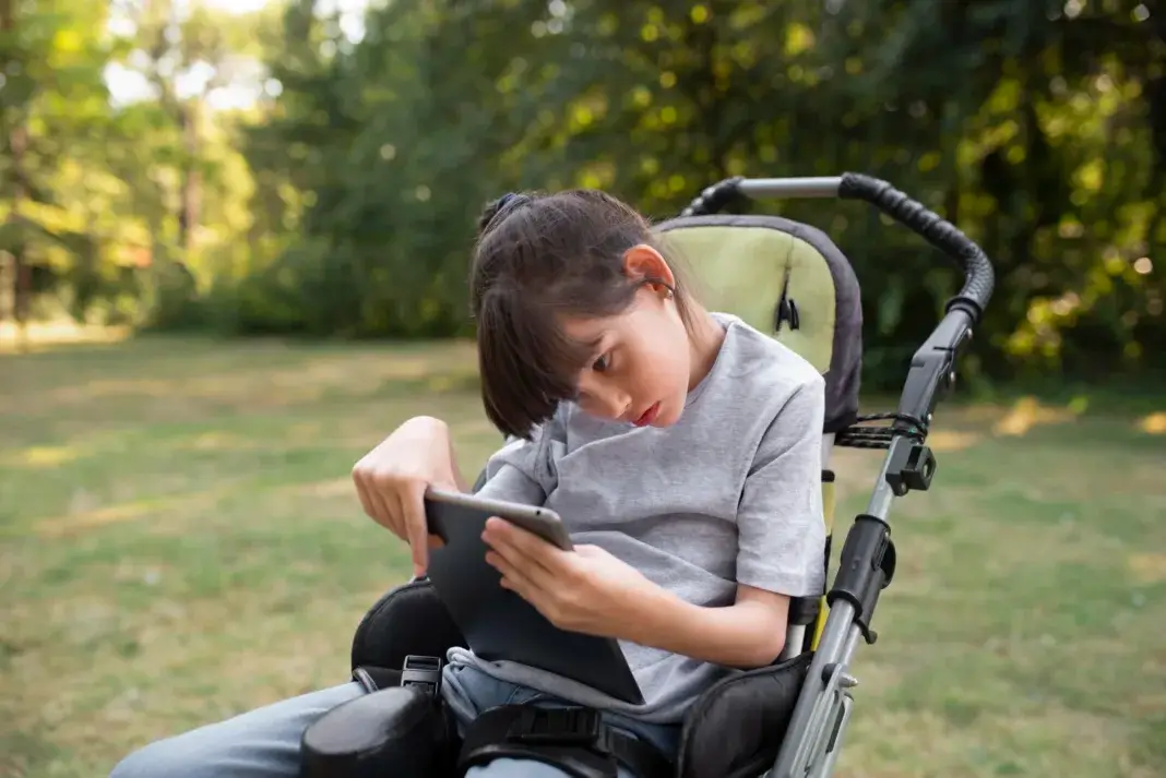 Enfant en situation de handicap, assis dans un fauteuil roulant, utilisant une tablette numérique en extérieur dans un parc verdoyant.