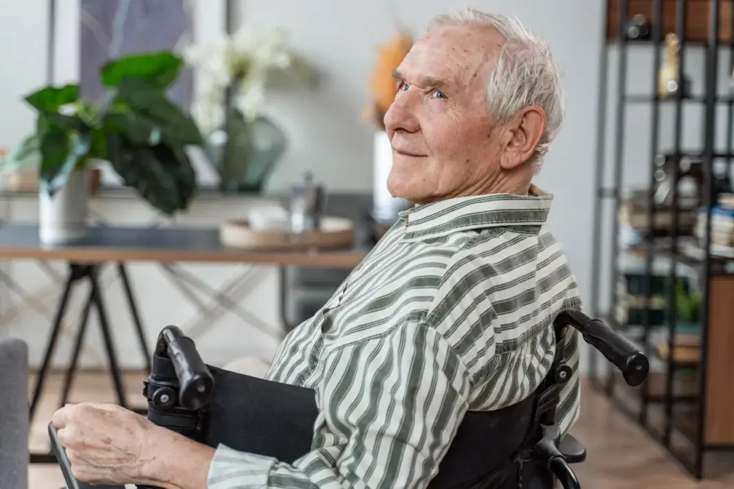 Personne âgée assise dans un fauteuil roulant à domicile, regard tourné vers l’extérieur, dans un environnement intérieur lumineux et apaisé