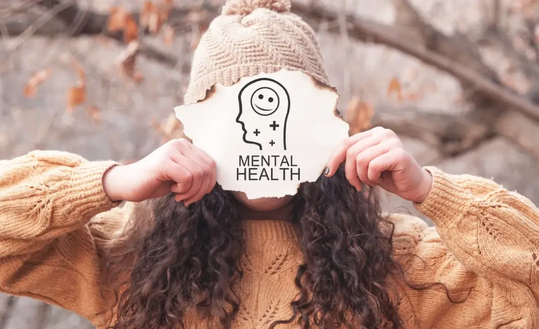 Personne tenant devant son visage une feuille découpée en forme de tête, illustrée d’un pictogramme de santé mentale avec un visage souriant et la mention « Mental Health »