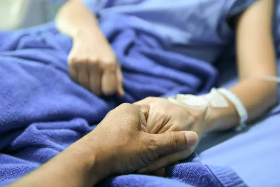 Main d’un proche tenant celle d’une personne allongée dans un lit médicalisé, évoquant l’accompagnement en fin de vie, la présence auprès du patient et le soutien apporté dans le cadre des soins palliatifs.