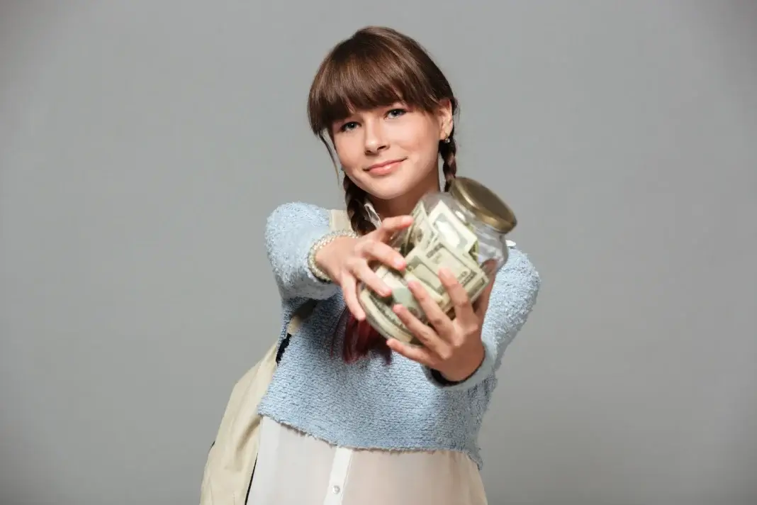 Jeune fille souriante présentant une tirelire en verre remplie de billets de banque, illustrant le soutien financier destiné aux jeunes en situation de précarité.