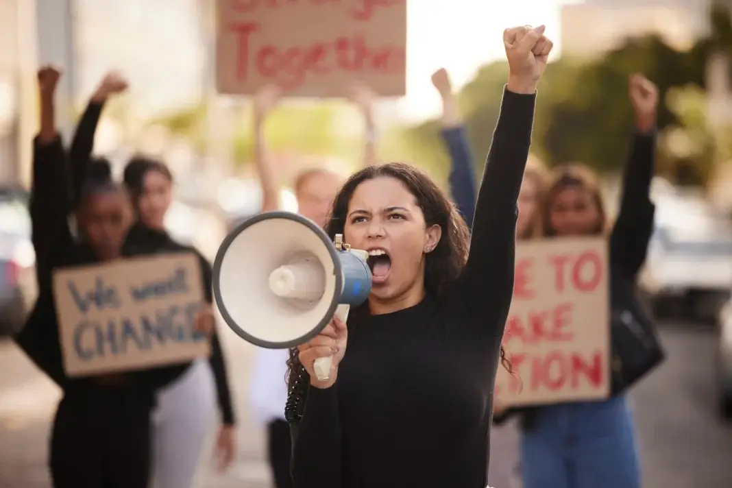 Une jeune femme manifeste dans la rue, un mégaphone à la main et le poing levé, entourée d’autres manifestants brandissant des pancartes. Une illustration des mobilisations collectives évoquées dans l’article consacré à l’actualité de la protection judiciaire de la jeunesse (PJJ) et au projet de loi « casseurs-payeurs ».